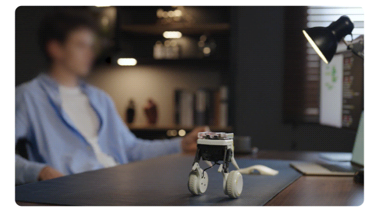 Small wheeled robot on a desk in a dim office, with a person blurred in the background.