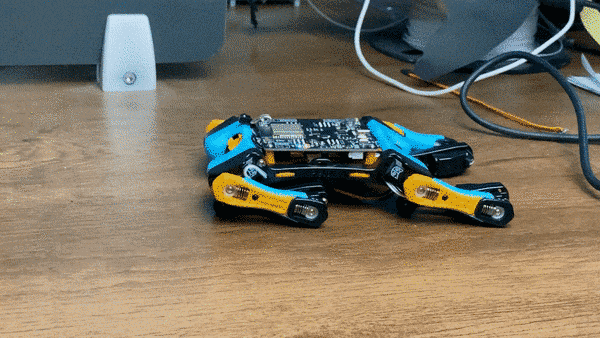 Small blue-and-yellow robotic chassis lying on a wooden desk with cables in the background.