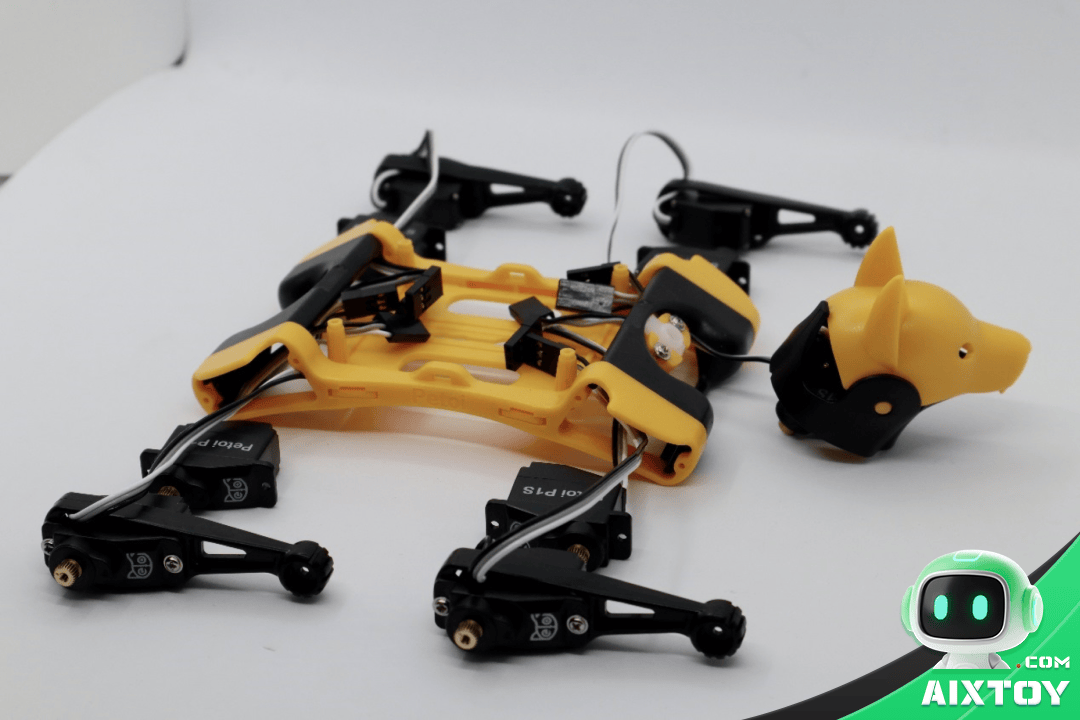 Yellow robotic chassis with four black leg modules and exposed wiring on a white surface; a yellow pig‑head accessory sits nearby on the right.