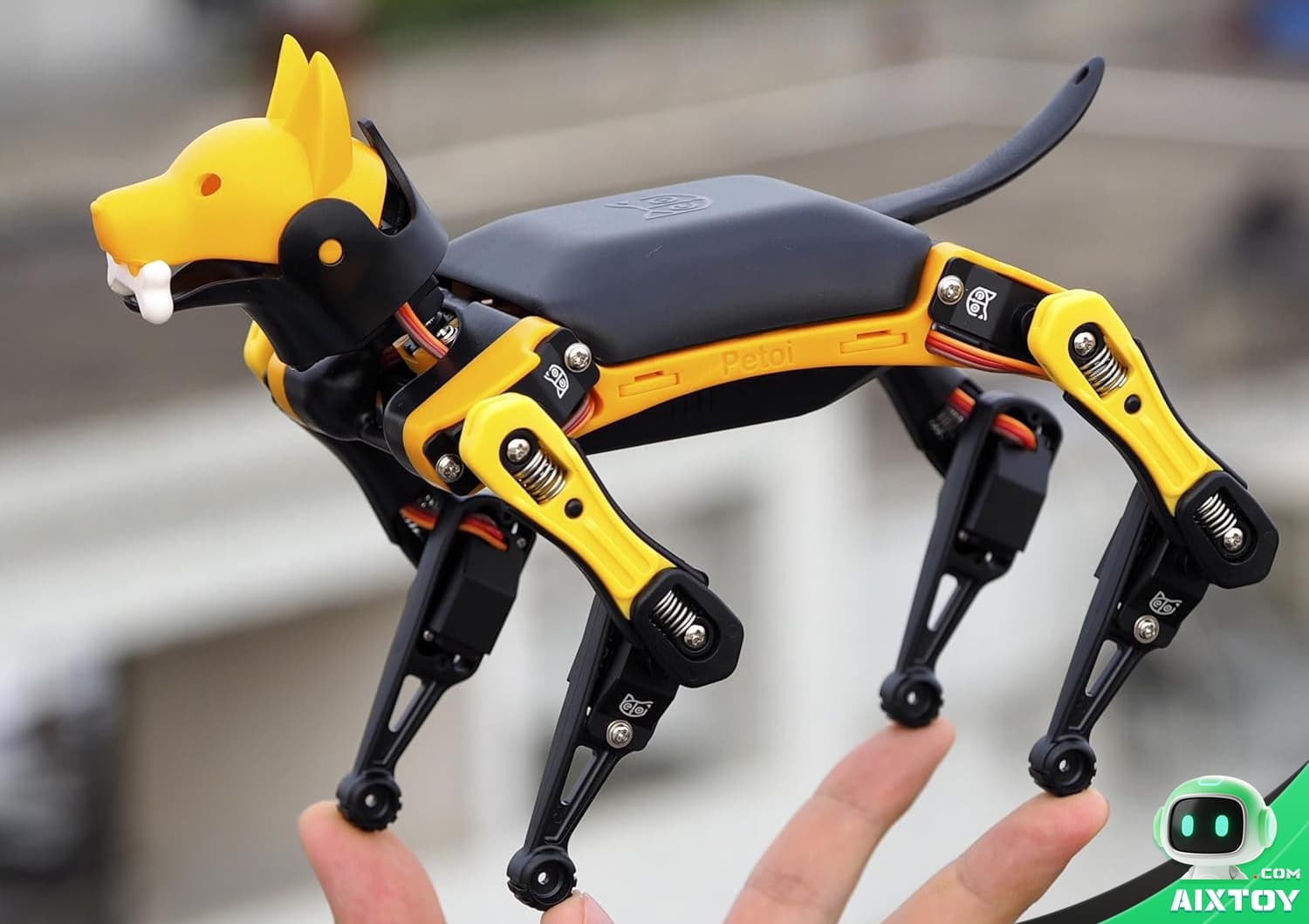 Yellow and black quadruped robot dog perched on a person's fingers, showing exposed joints and mechanical actuators on its legs.