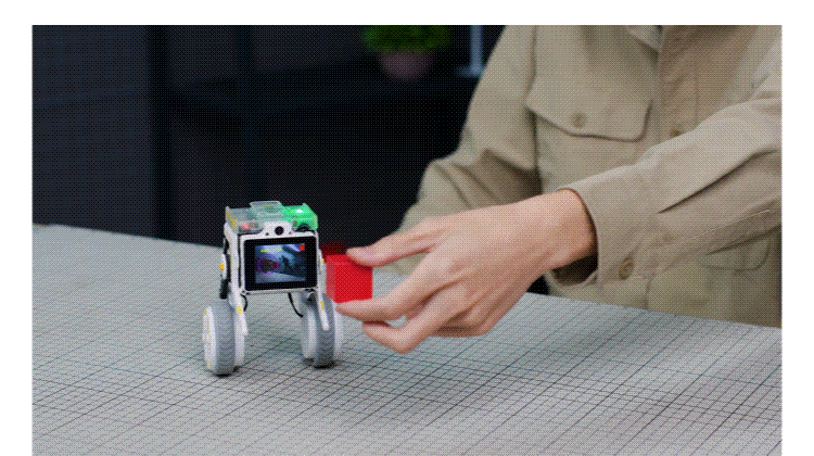 Small four-wheeled educational robot with a front screen on a grid tabletop as a person places a red cube near it.