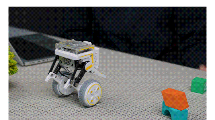 Small wheeled educational robot on a grid-pattern table with colorful blocks nearby.