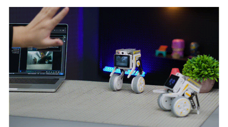Person's hand reaches toward a laptop on a desk with two small wheeled robots nearby and a potted plant in the background.