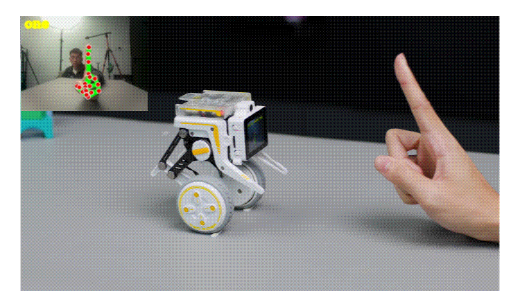 Small white educational robot on a gray table, with a hand pointing toward it, implying interaction; inset video shows a person observing.