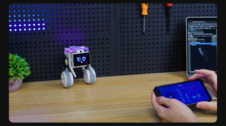 Small wheeled robot on a wooden desk facing a person holding a blue-screen smartphone, with a monitor showing code in the background.