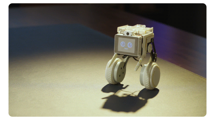 Small white wheeled robot with a screen face standing on a pale surface, casting a shadow nearby.
