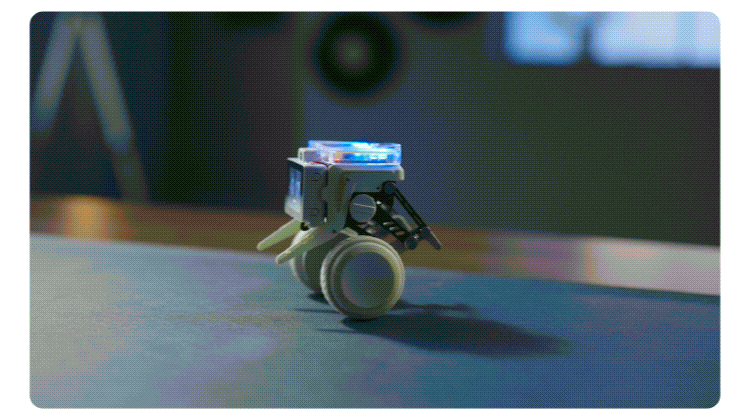 Small wheeled robot prototype on a dark tabletop with blue-lit components on top in a lab setting.