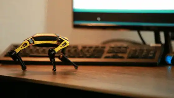 Animated demo of a yellow and black Petoi robot dog walking across a desk in front of a monitor, running the open-source OpenCat gait engine